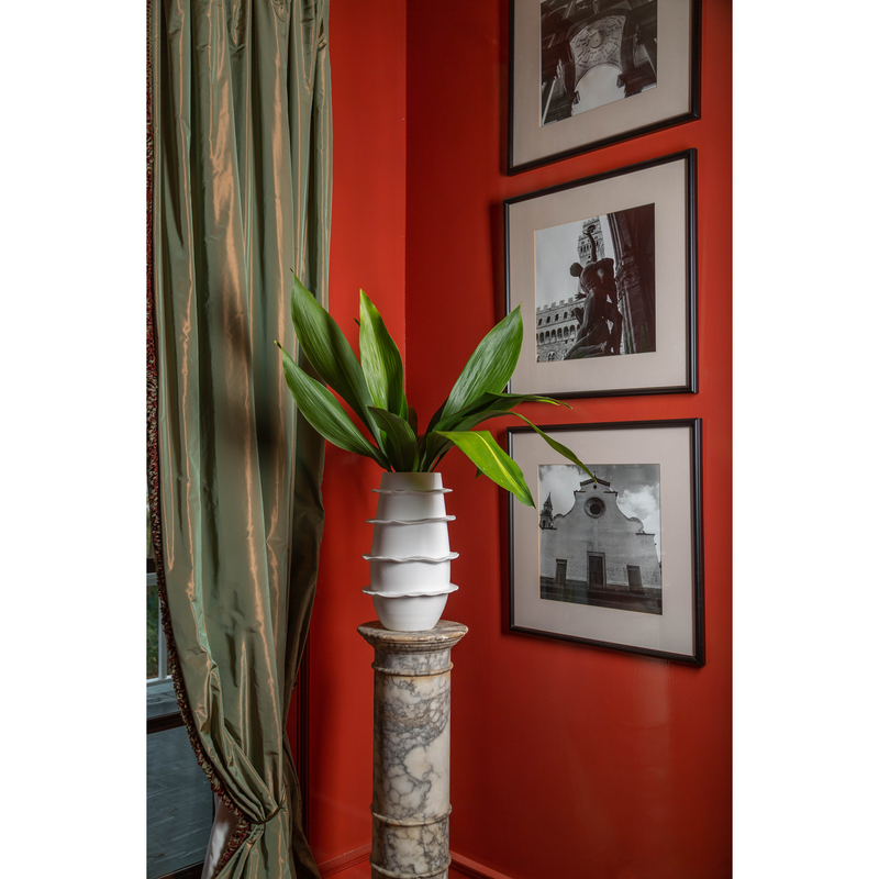 Galaxy Spiral Matte Tall Vase in white holds fresh green leaves, set on a marble pedestal. The red wall behind is adorned with black and white photos, complemented by green curtains. Handmade in Italy by Abigails, this ceramic piece adds an elegant touch to any decor.