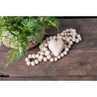 The Large Heart Wooden Prayer Beads from Sugarboo & Co. rest gracefully on a rustic wooden surface, accompanied by a small woven basket that holds a vibrant green fern plant, creating a distinctive home accent.
