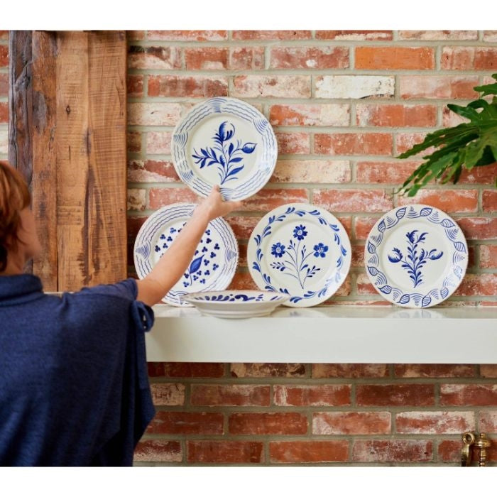 A person arranges Abigails Casa Nuno Blue Flower Dinner Plates (set of 2) on a white shelf against a brick wall.