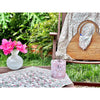 A wooden chair featuring a floral cloth and a wicker bag, adorned with pink flowers in a white vase, and accompanied by a pink candle in a garden setting, beautifully complemented by Abigails' Pomegranate Cabana Napkins (set of 4).