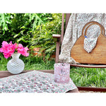 A wooden chair featuring a floral cloth and a wicker bag, adorned with pink flowers in a white vase, and accompanied by a pink candle in a garden setting, beautifully complemented by Abigails' Pomegranate Cabana Napkins (set of 4).