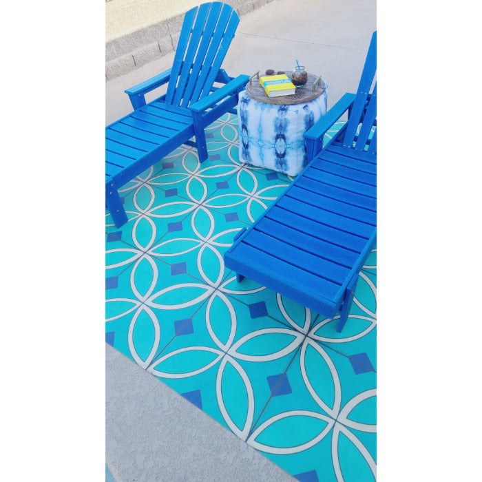 Two blue Adirondack chairs on a Pattern 70 Echoes From The Bells vinyl floorcloth by a small table with books and a candle.