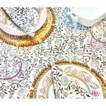 A table setting featuring floral-patterned plates, woven placemats, and clear glasses rests on a floral tablecloth, enhanced by Abigails' Pomegranate Cabana Napkins (set of 4) for a touch of elegance.