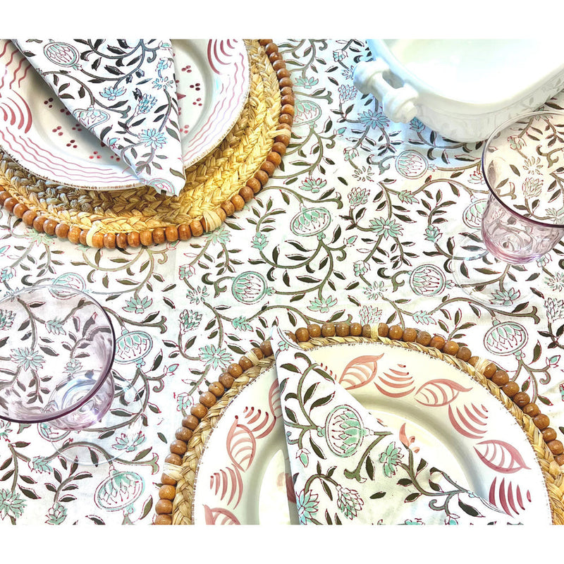 A table setting featuring floral-patterned plates, woven placemats, and clear glasses rests on a floral tablecloth, enhanced by Abigails' Pomegranate Cabana Napkins (set of 4) for a touch of elegance.