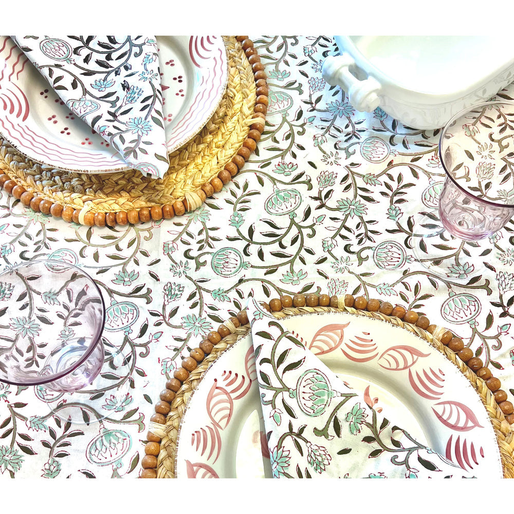 The table is set with Majolica ceramics, folded napkins, woven placemats, and pink glasses on a floral tablecloth. Casa Nuno Pink Dinner Plates by Abigails add a touch of elegance to the arrangement.