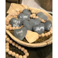 The Large Heart Wooden Prayer Beads from Sugarboo & Co. include a wooden bowl filled with heart-shaped stones inscribed with messages such as "LOVE IS love," "JE T'AIME," "Always," "loved," and "xoxo." This distinctive home accessory is adorned with oversized wooden beads and moss.