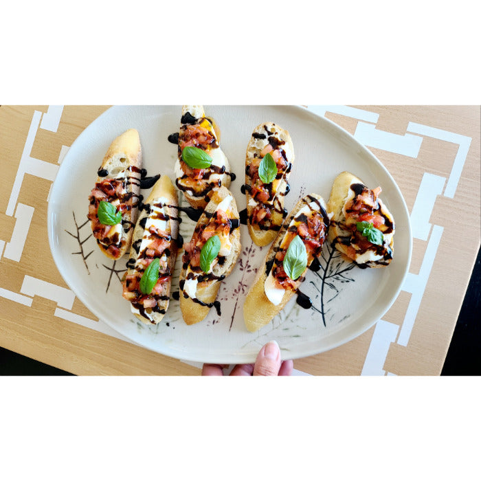 A plate of bruschetta topped with tomato, basil, and balsamic glaze rests elegantly on a COCOCOZY Square Link Large Serving Board by BSEID, enhancing the rustic charm against the wooden table.