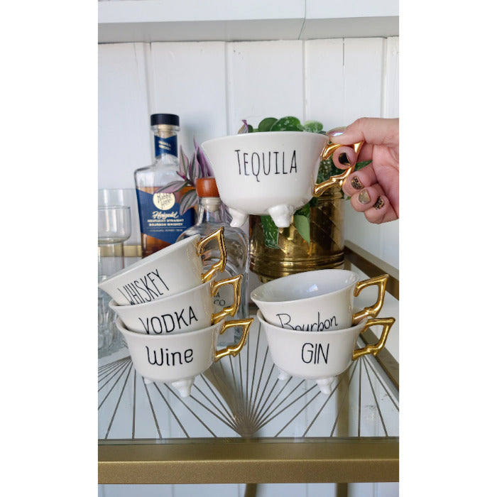 Stacked Creative Co-Op's White + Gold Stoneware Footed Tea Cups (set of 6), labeled with alcohol names and featuring gold handles, sit on a glass table. A hand holds the "Tequila" serving cup—perfect bar decor.