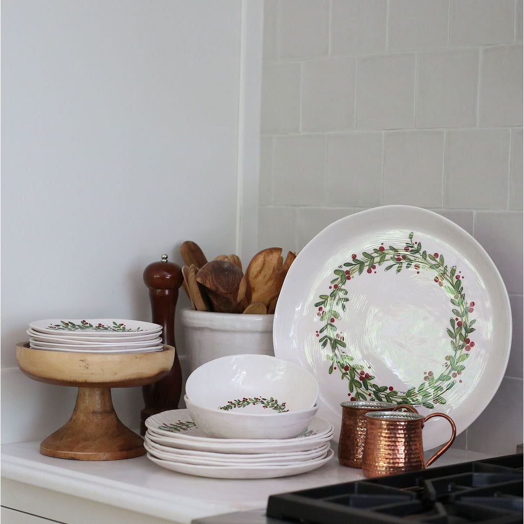 Floral white plates stacked beside Always Relish's Christmas Wreath Melamine Serving Bowl and wooden utensils on the counter.