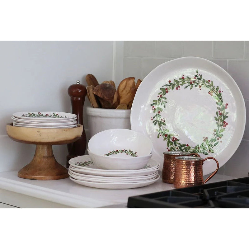 Stacked white ceramic plates with a green and red wreath design sit alongside a copper mug, a wooden pepper mill, and wooden utensils on a kitchen counter. A set of Always Relish Christmas Wreath Melamine Serving Platters adds an extra festive touch to the scene.