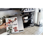 A hand holds a cookbook titled "Market Math" in a kitchen with a BSEID Stainless Recipe Book / iPad Holder and a roll of paper towels.