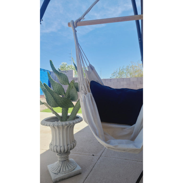 The Hand Loomed Hanging Hammock Chair from Under The Bough, featuring a black cushion, sits gracefully next to a large cactus in an ornate pot on a patio with a clear sky backdrop. This exquisite piece, handmade in Paraguay from 100% cotton, adds both comfort and elegance to your outdoor space.
