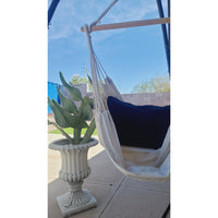 The Hand Loomed Hanging Hammock Chair from Under The Bough, featuring a black cushion, sits gracefully next to a large cactus in an ornate pot on a patio with a clear sky backdrop. This exquisite piece, handmade in Paraguay from 100% cotton, adds both comfort and elegance to your outdoor space.