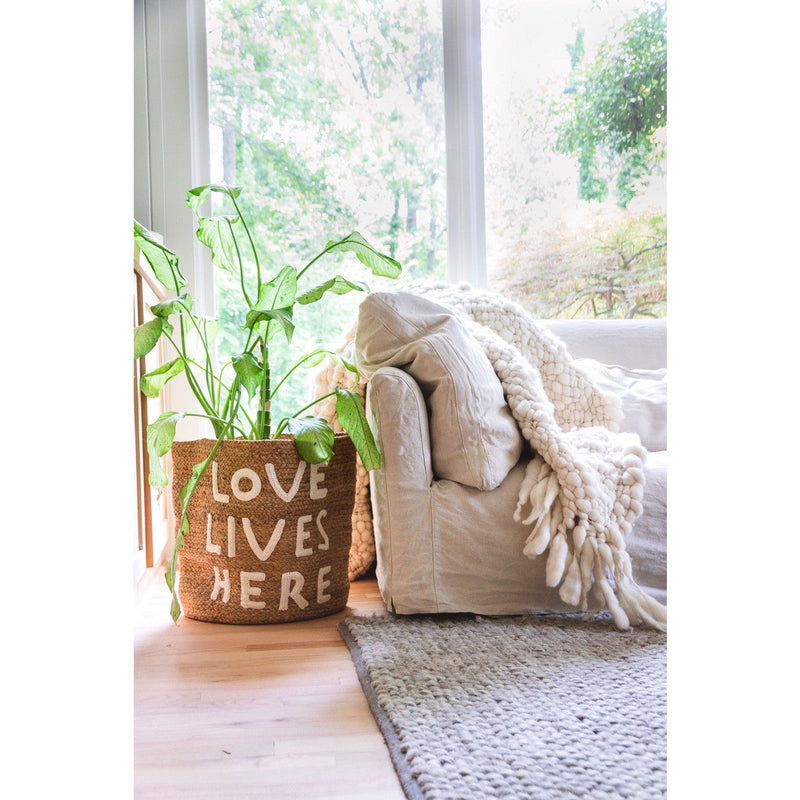 A cozy living room features the Sugarboo & Co. "Love Lives Here" Jute Basket and a sofa adorned with a knitted blanket, adding a perfect textural accent.