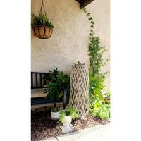 Outdoor scene with a black bench, a hanging planter, tall woven lantern in driftwood tone, and various potted plants against a stucco wall. Kalalou's Willow Extra Large Glass Candle Lanterns and LED battery-operated candles add a cozy ambiance.