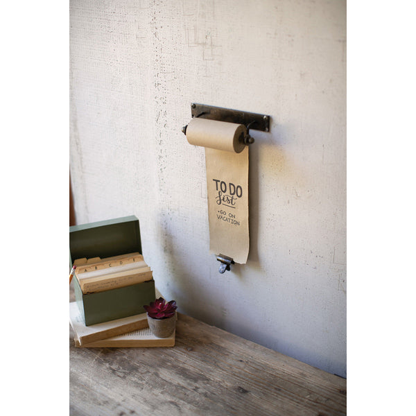 A wall-mounted Kalalou Hanging Note Roll (7") with "TO DO list: go on vacation" written on it, next to a desk adorned with books, a plant, and an antique brass clip.