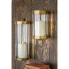 Two Kalalou Glass + Antique Brass Candle Wall Sconces with white candles adorn a rustic wooden wall beside books.