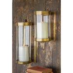 Two Kalalou Glass + Antique Brass Candle Wall Sconces with white candles adorn a rustic wooden wall beside books.