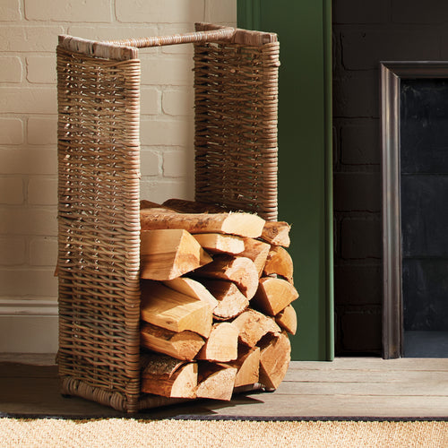Near the fireplace, a Napa Home & Garden Rattan Log Basket - Tall sits on the wooden floor, filled with firewood.