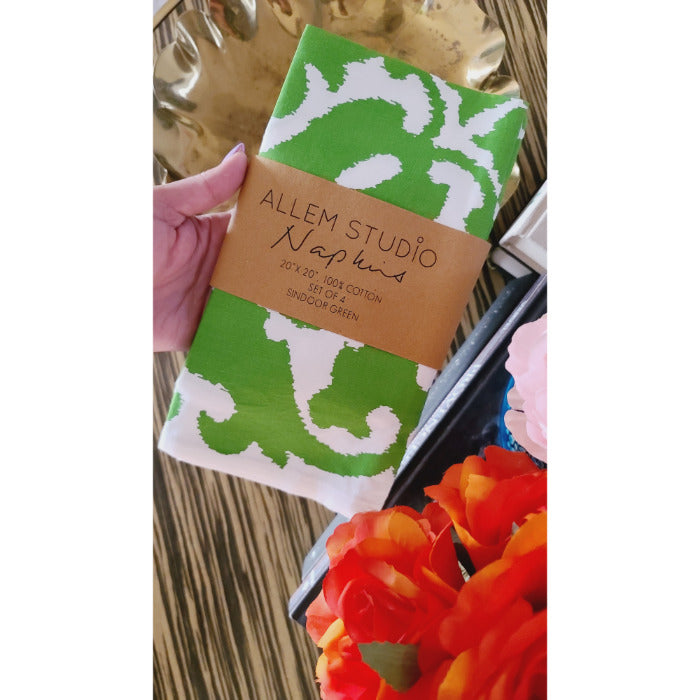 A hand holds a Sindoor Green Screened Napkin from Allem Studio, featuring a green and white damask pattern, near colorful flowers.