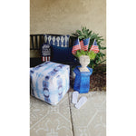 Patio scene with a fade-resistant blue pouf, Newbury Velvet Lumbar by Lacefield, U.S. flags, plant, and white flip-flops.