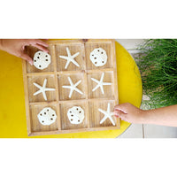 Two hands enjoy the Kalalou Tic-Tac-Toe Seaside Game, playing with sand dollars and starfish as pieces on a yellow table.