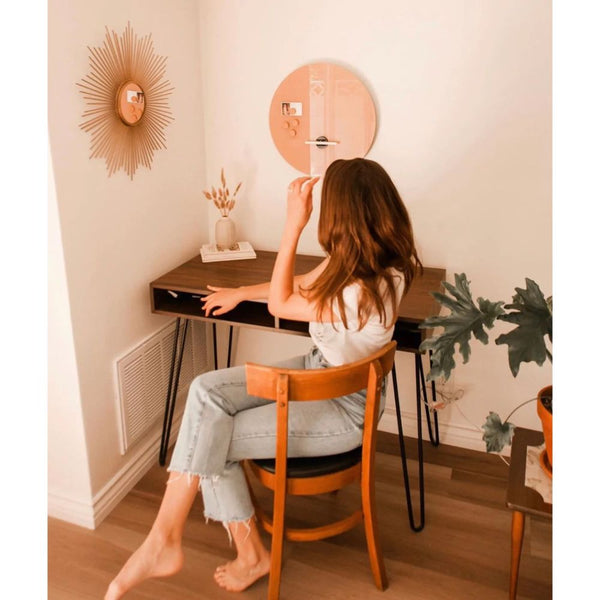 A person sits at a desk, touching their hair, surrounded by plants and accentuated by a sunburst mirror on the wall, while the Glass Magnetic Dry Erase Round Board - Clay from Petal Lane Home enhances the contemporary aesthetic of the room.