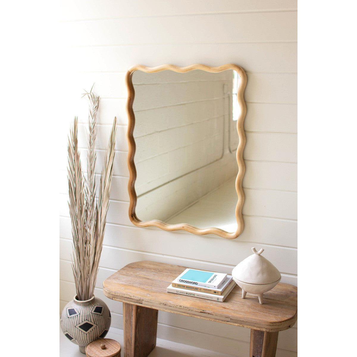 The Kalalou Wooden Squiggle Wall Mirror, complete with hanging hardware, adorns a white paneled wall above a wooden bench that holds two books and a small beige decorative object. To the left of the bench, an intricately patterned vase contains tall dried grasses, enhancing the minimal and rustic setting.