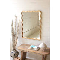 The Kalalou Wooden Squiggle Wall Mirror, complete with hanging hardware, adorns a white paneled wall above a wooden bench that holds two books and a small beige decorative object. To the left of the bench, an intricately patterned vase contains tall dried grasses, enhancing the minimal and rustic setting.