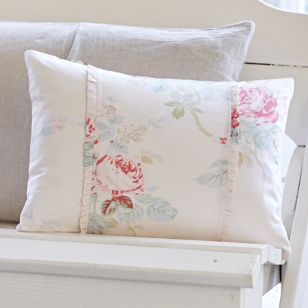 A pillow from the Shore Rose Petal Bedding Collection by Taylor Linens, featuring a floral pattern and ruffles, sits on a light grey cushion in a white seat setting, adding a charming touch.