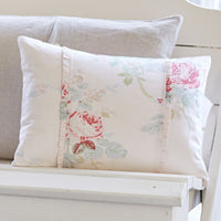 A pillow from the Shore Rose Petal Bedding Collection by Taylor Linens, featuring a floral pattern and ruffles, sits on a light grey cushion in a white seat setting, adding a charming touch.